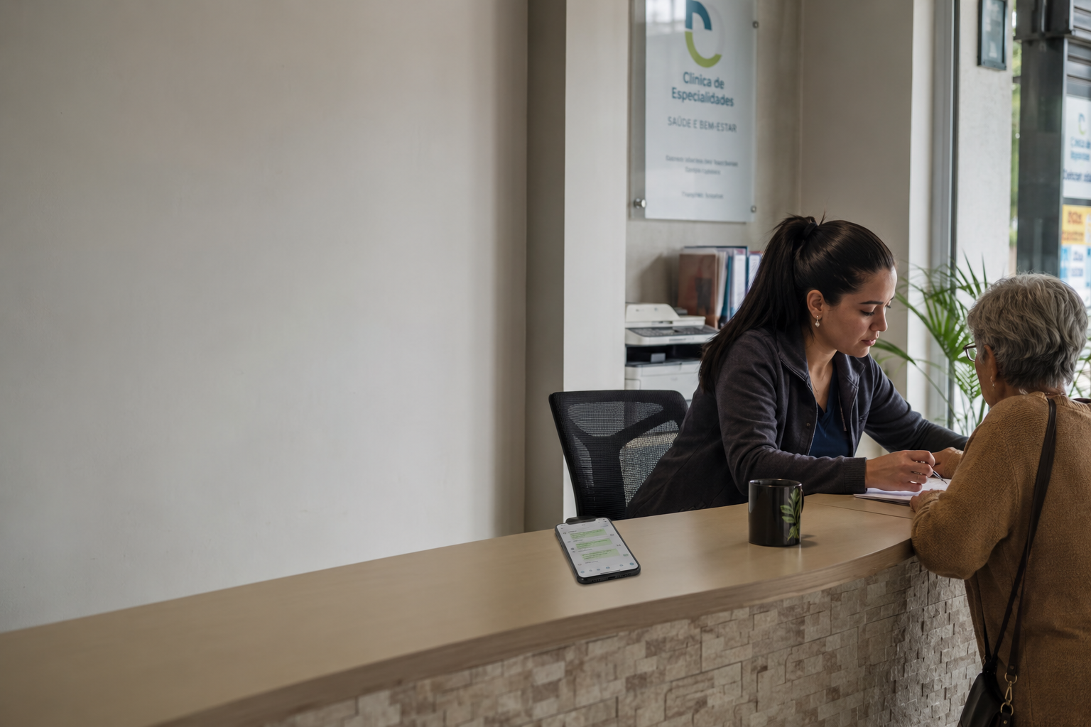 Uma recepcionista de clínica atendendo pessoalmente uma paciente idosa no balcão, enquanto o celular ao lado mostra conversas de WhatsApp sendo resolvidas pelo agente.
